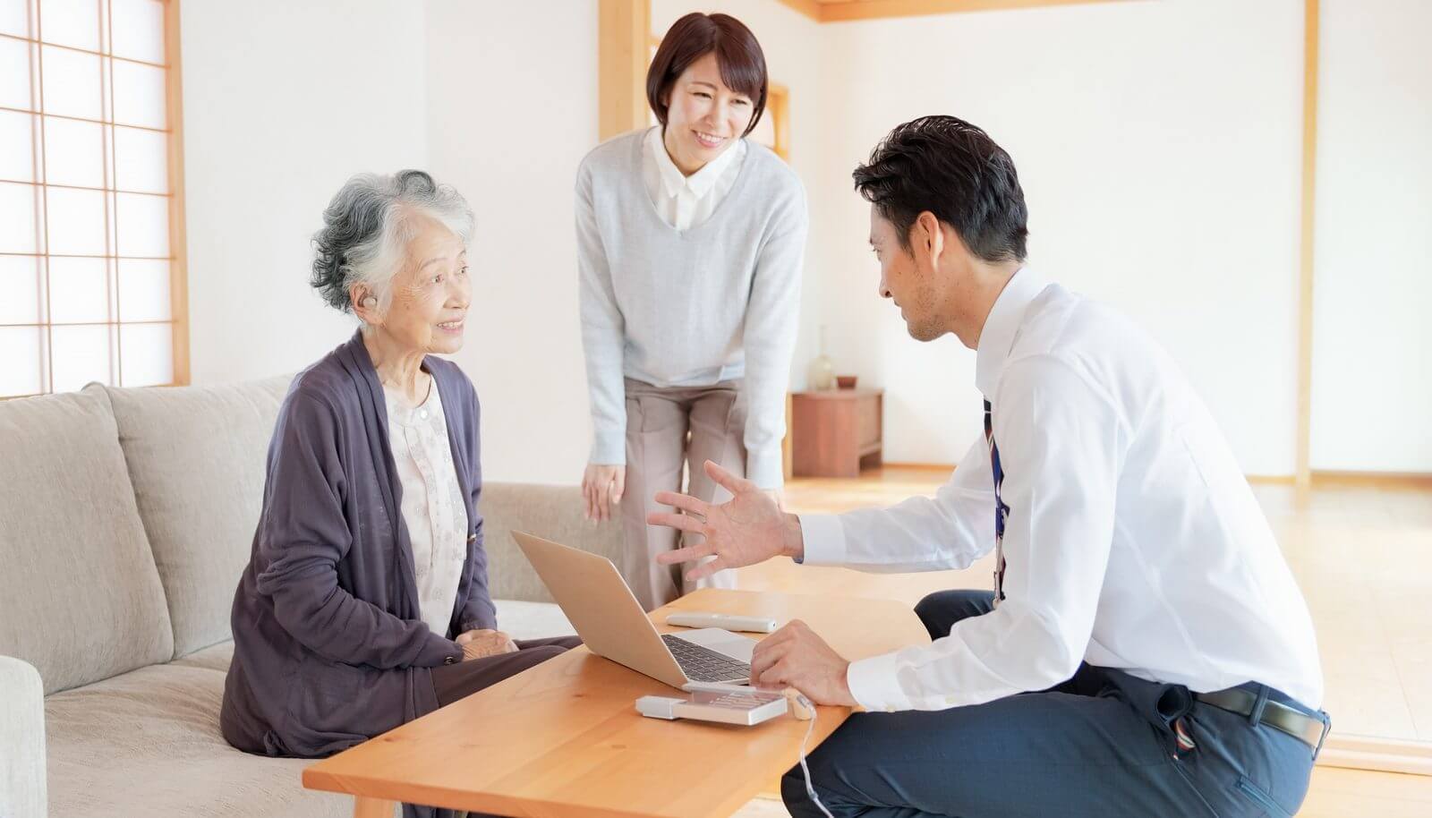 自宅を訪問した補聴器のスタッフが、測定機器とパソコンを使って高齢の女性と家族に説明している様子